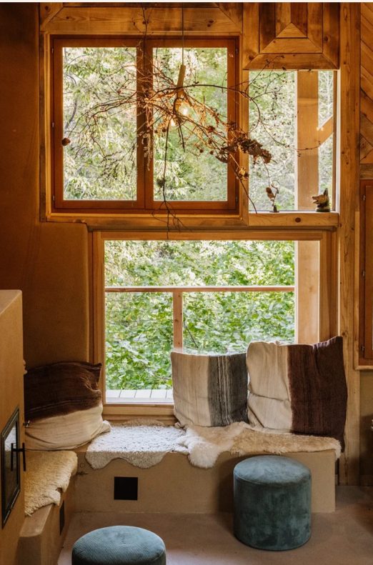 Zitbank met kussens voor een groot houten raam met zicht op het bos in gîte Le Sapin du Renard