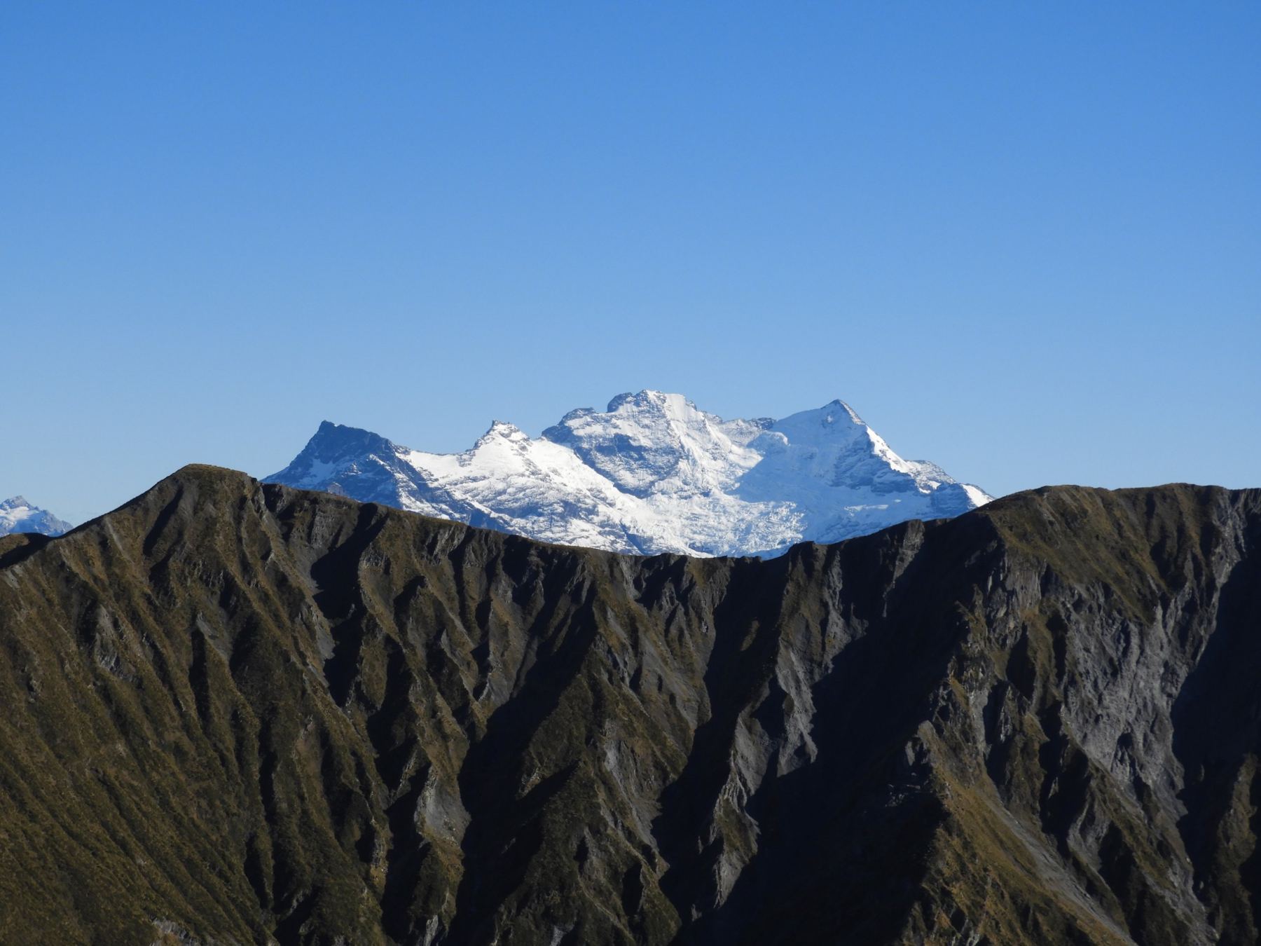 Besneeuwde bergtop achter een donkere bergkam onder een strakblauwe lucht